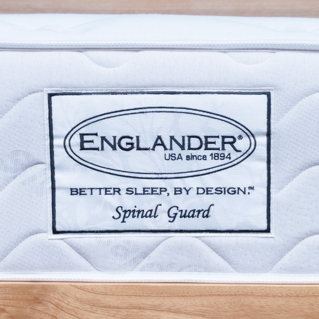 A close-up of a white quilted Englanders Spinal Guard mattress or bedding surface with a sewn-on label reading ENGLANDER, USA since 1894, Better Sleep, by Design, and Spinal Guard, set against a light wood background.