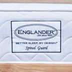 A close-up of a white quilted Englanders Spinal Guard mattress or bedding surface with a sewn-on label reading ENGLANDER, USA since 1894, Better Sleep, by Design, and Spinal Guard, set against a light wood background.