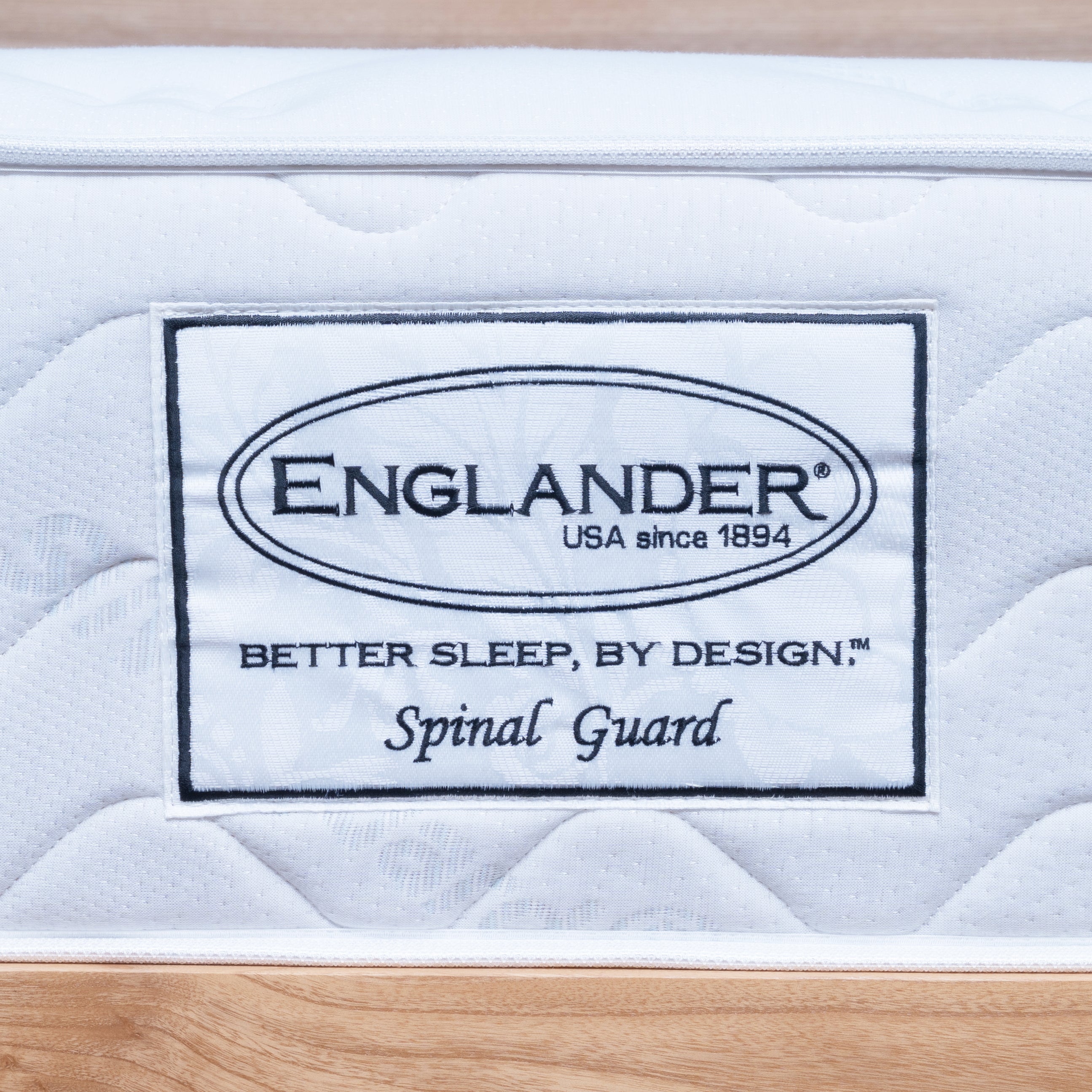 A close-up of a white quilted Englanders Spinal Guard mattress or bedding surface with a sewn-on label reading ENGLANDER, USA since 1894, Better Sleep, by Design, and Spinal Guard, set against a light wood background.