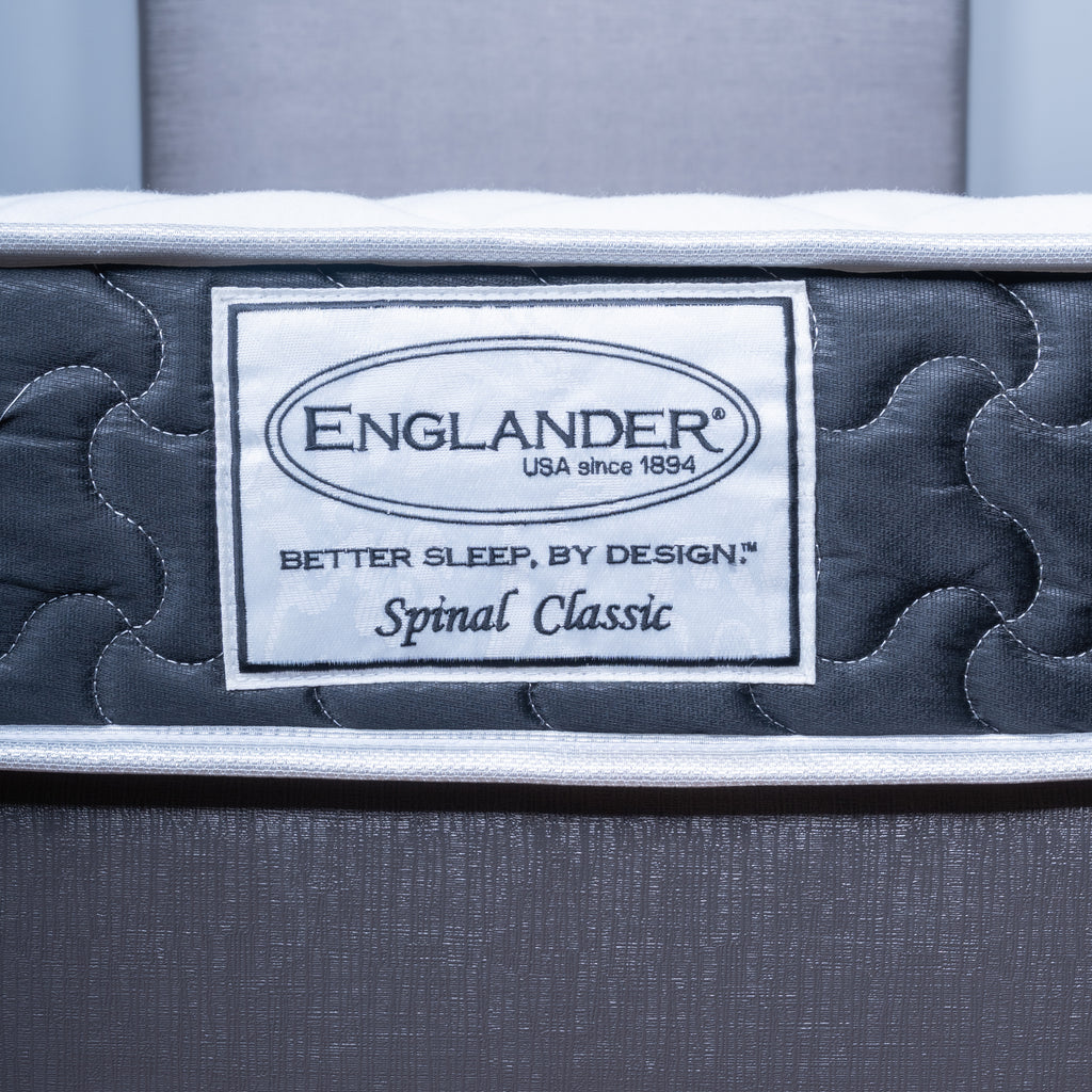 Close-up of an Englander Spinal Classic mattress showing the brand label on the side, with dark gray quilted padding and a white border.
