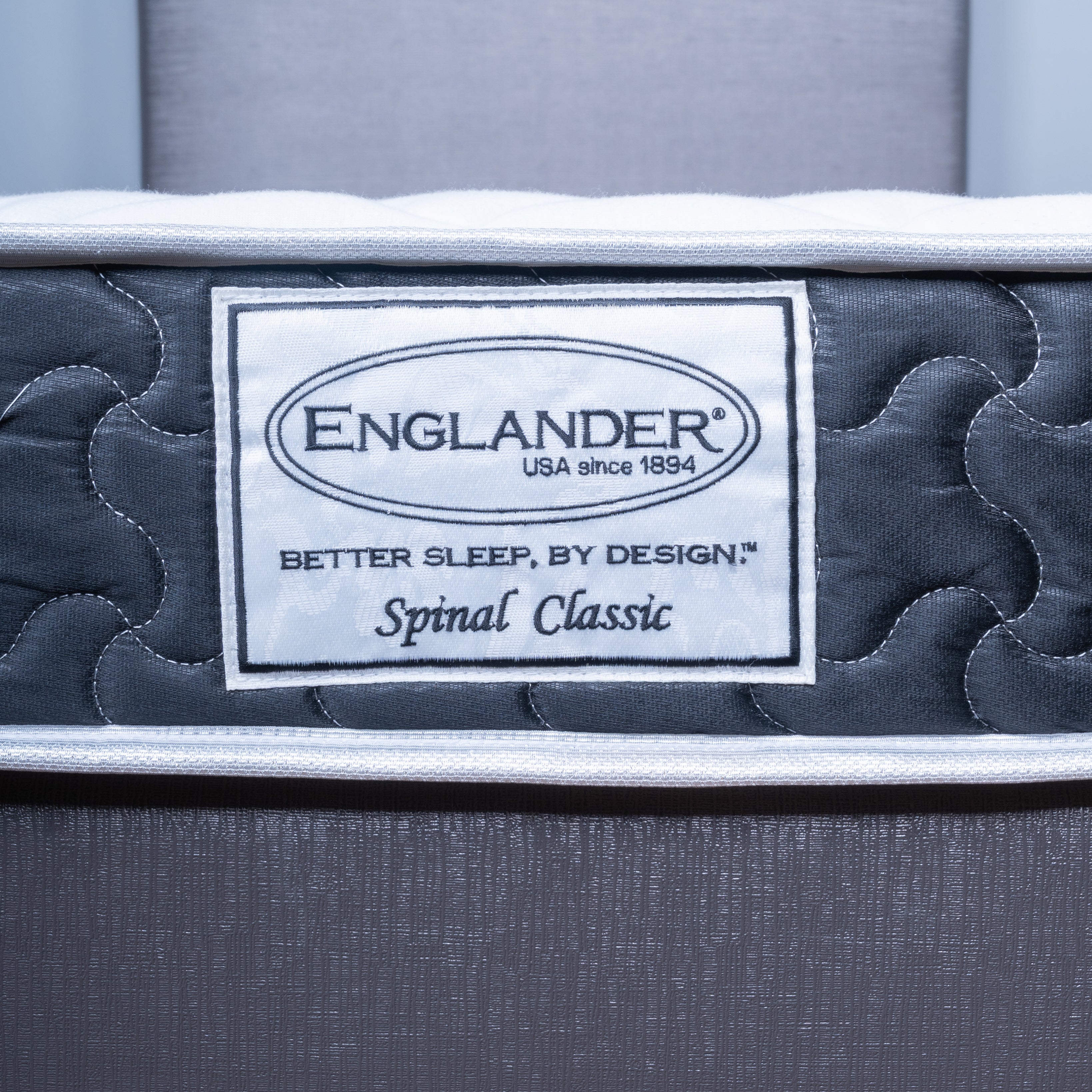 Close-up of an Englander Spinal Classic mattress showing the brand label on the side, with dark gray quilted padding and a white border.