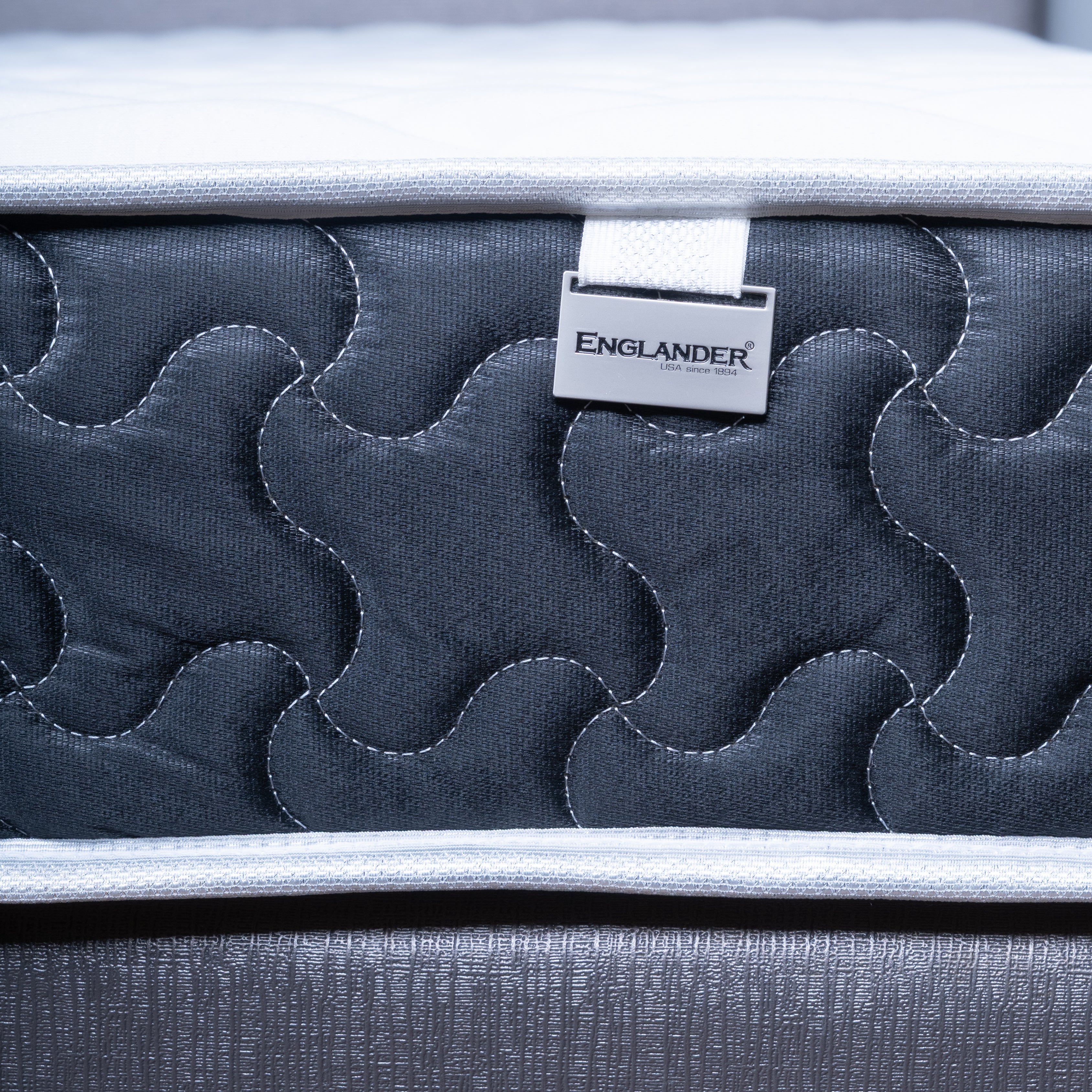 Close-up of a Englanders Spinal Classic mattress side panel with a visible ENGLANDER brand tag, showing dark gray quilted stitching and a light gray fabric edge.