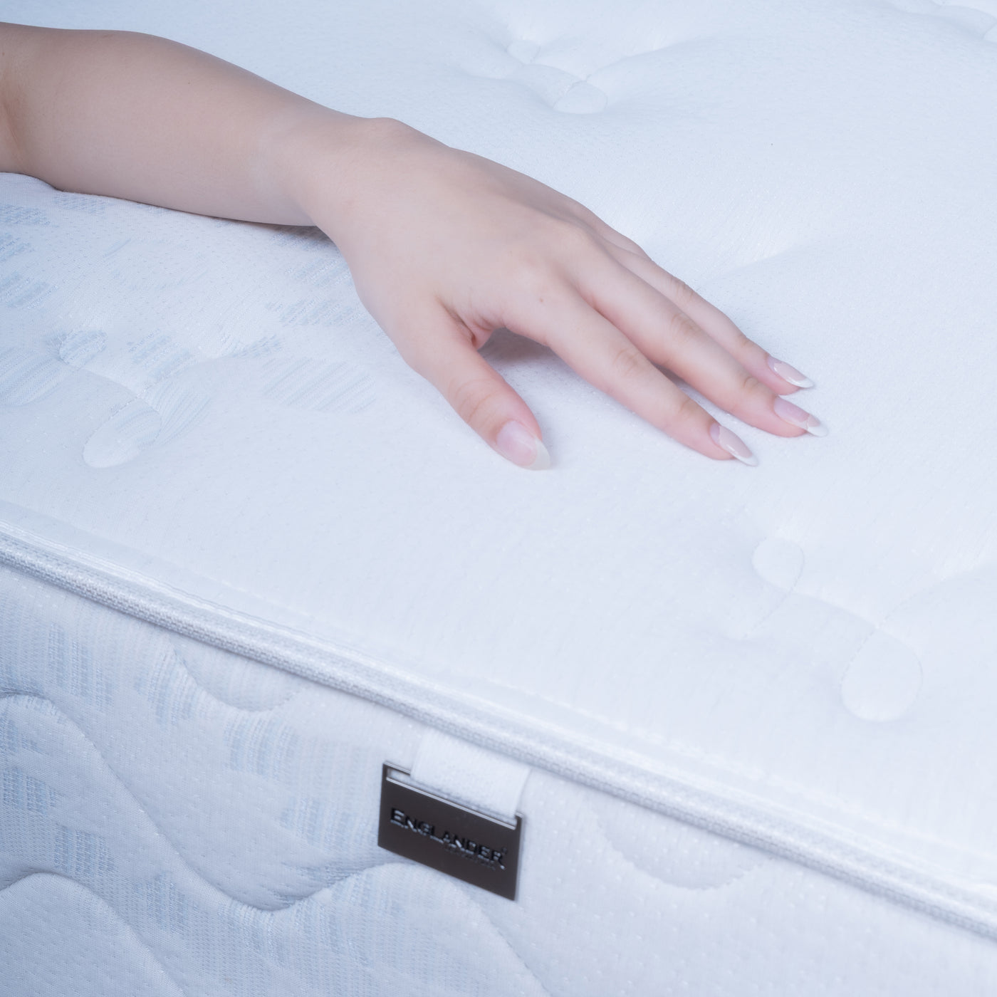 A close-up of a person's hand resting on a white quilted Spinal Guard Mattress, with a small black brand tag visible near the edge.