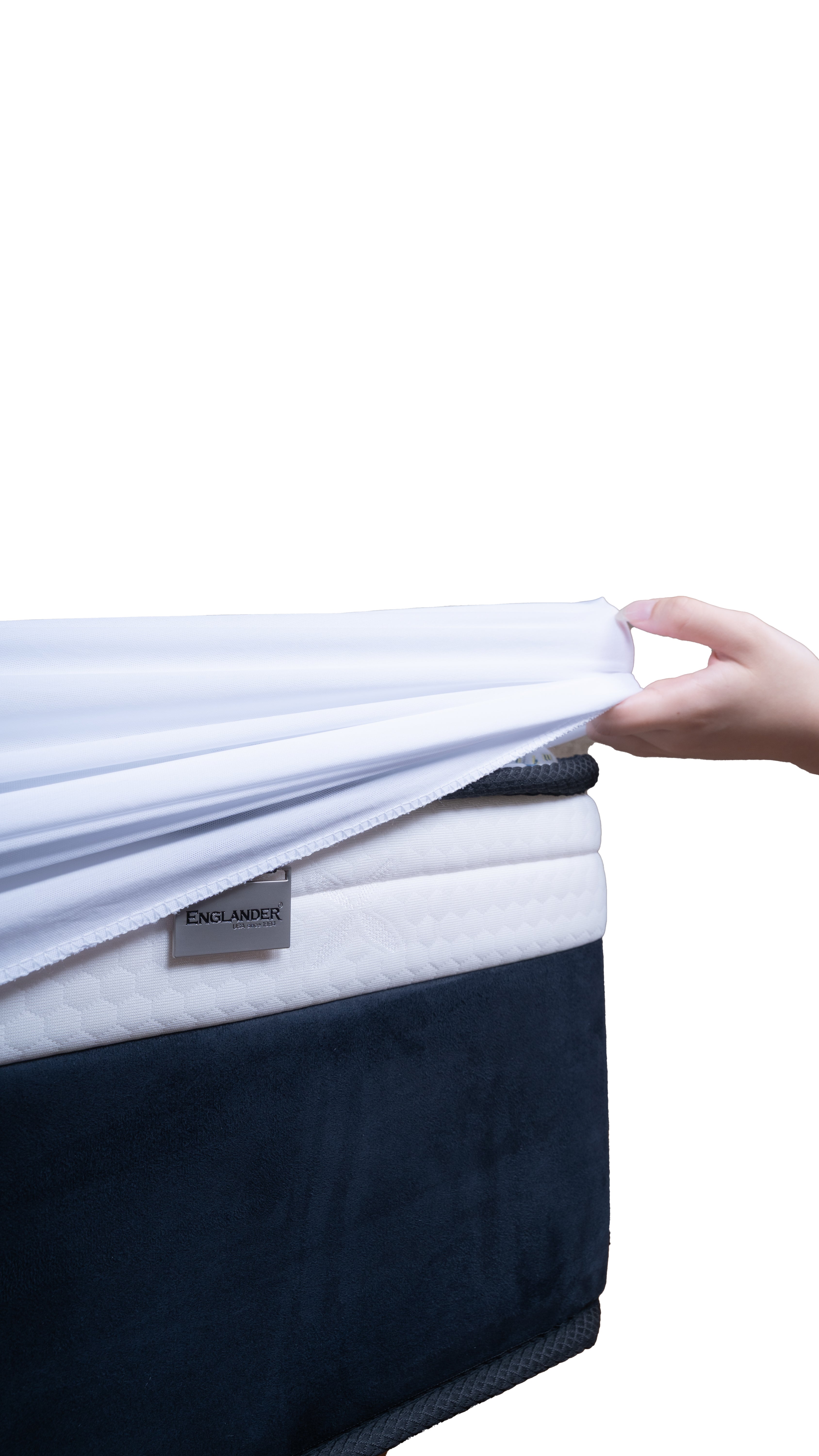 A close-up of a mattress with a white sheet being pulled upward by a hand, showing the mattress layers and a dark navy blanket or cover on the lower portion. A small tag reading ENGLANDER is visible on the mattress.